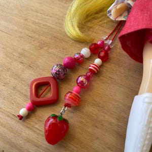 May include: A close-up of a doll's accessories, including a red square pendant and a beaded necklace with pink, red, and white beads. The necklace features a red strawberry charm. The doll is wearing a red dress and white boots.
