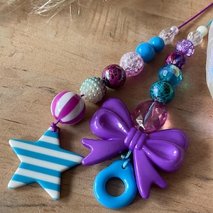 May include: A close-up of two beaded accessories. One features a purple bow and a blue ring, the other a star with blue and white stripes. Both have colorful beads in shades of purple, blue, and white.