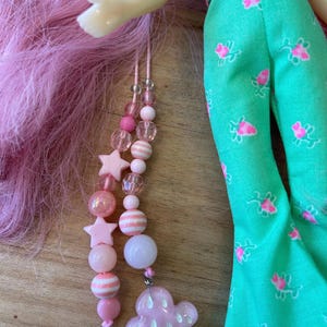 May include: A pink beaded accessory with star and round beads, and a cloud-shaped charm. The accessory is hanging near a doll with pink hair and a green jumpsuit with pink floral patterns. The jumpsuit has a yellow belt.