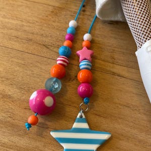 May include: A colorful beaded necklace with a star pendant. The necklace features a variety of beads in pink, orange, blue, and white. The star pendant is white with blue stripes. The necklace is on a wooden surface.