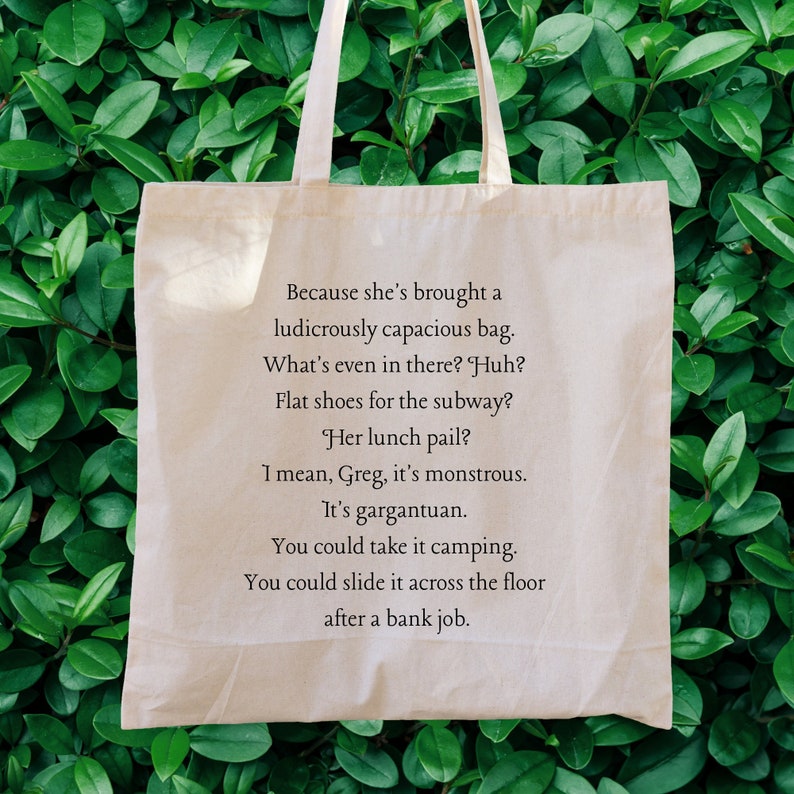 May include: A beige tote bag with a quote printed on it. The quote reads: "Because she's brought a ludicrously capacious bag. What's even in there? Huh? Flat shoes for the subway? Her lunch pail? I mean, Greg, it's monstrous. It's gargantuan. You could take it camping. You could slide it across the floor after a bank job."
