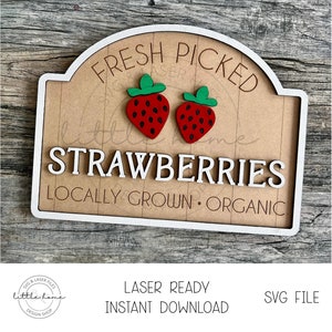 Könnte beinhalten: Ein Schild aus Holz mit weißem Rand und braunem Hintergrund. Das Schild trägt die Aufschrift "Frisch gepflückte Erdbeeren Lokal angebaut Bio". Auf dem Schild befinden sich zwei rote Erdbeeren mit grünen Blättern.