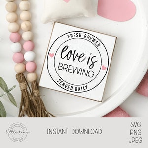 Op de afbeelding: Een wit houten bord met een zwarte cirkel en de tekst "Fresh Brewed Love is Brewing Served Daily" met twee roze hartjes. Het bord ligt op een wit bord met een roze hartje en een houten kralensnoer met roze en witte kralen.