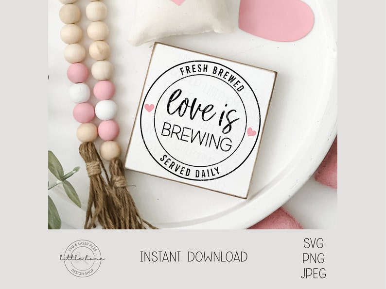 Op de afbeelding: Een wit houten bord met een zwarte cirkel en de tekst "Fresh Brewed Love is Brewing Served Daily" met twee roze hartjes. Het bord ligt op een wit bord met een roze hartje en een houten kralensnoer met roze en witte kralen.