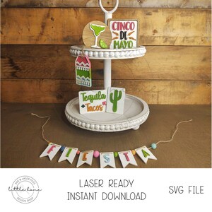 May include: A two-tiered white tiered tray with a green cactus, a green margarita glass, a pink and green tag, and a sign that says "Tequila & Tacos". The top tier has a sign that says "Cinco de Mayo". A banner with the word "FIESTA" is in the bottom of the image.