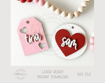 May include: Two wooden Valentine's Day ornaments with hearts and names. The first ornament is pink with a white heart cutout and the name "tina" in white. The second ornament is white with a red heart and the name "sam" in white.