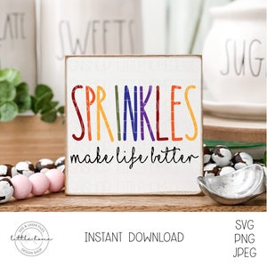 Könnte beinhalten: Ein weißes Holzschild mit dem Text "SPRINKLES make life better" in Regenbogenfarben. Das Schild steht auf einer Holzoberfläche mit einer rosa-weißen Perlenkette und einer silbernen Schaufel.