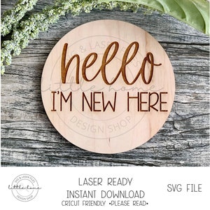 May include: A round wooden sign with the text "hello little home I'M NEW HERE" in brown lettering. The sign is laser cut and ready to be used for a new baby announcement.