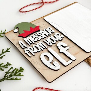 May include: A wooden sign with a white background and the text "A message from your elf" in black and white letters. The sign has a green and red elf hat on top.