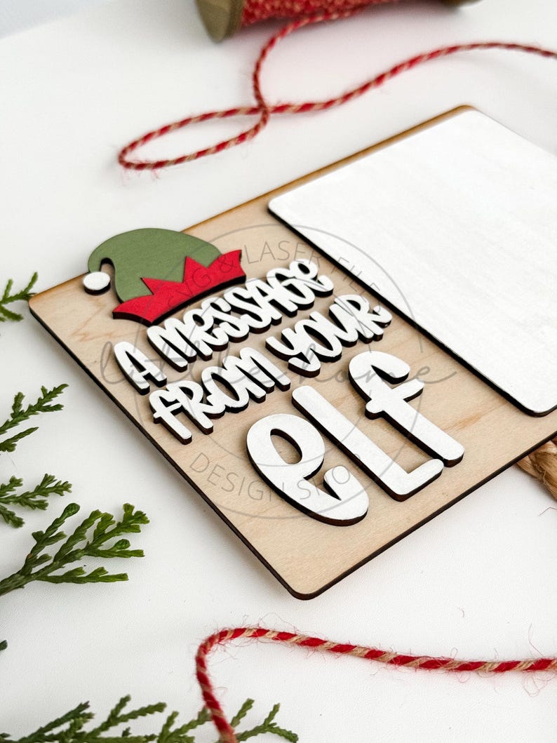 May include: A wooden sign with a white background and the text "A message from your elf" in black and white letters. The sign has a green and red elf hat on top.