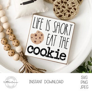 Puede incluir: Un letrero cuadrado de madera con el texto "LIFE IS SHORT EAT THE COOKIE" y un gráfico de una galleta con un mordisco. El letrero está sobre una bandeja blanca con cuentas de madera decorativas y una pequeña almohada.