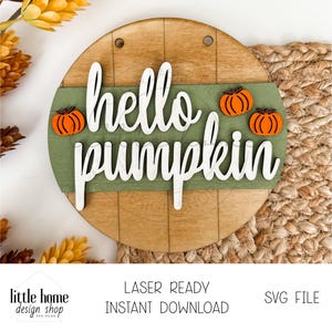 以下が含まれることがあります： 白い筆記体で「hello pumpkin」と書かれた木製の丸い看板。緑色のストライプがあり、オレンジ色のカボチャの切り抜きが3つあります。背景は明るい茶色の木目です。