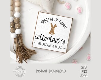 May include: A white wooden sign with the text "SPECIALTY CANDY cottontail co. -- JELLYBEANS & PEEPS --" in black lettering. A brown bunny silhouette is centered above the text. The sign is on a white surface with a brown and white beaded garland and pink paw print decorations.