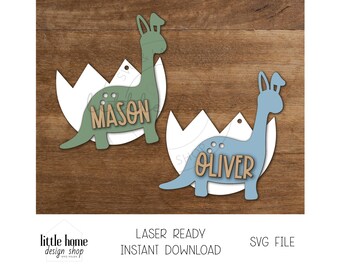 May include: Two dinosaur-shaped wooden cutouts with names "Mason" and "Oliver" inside egg-shaped cutouts. The dinosaurs are green and blue, and the eggs are white.
