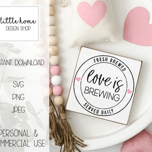 Op de afbeelding: Een wit houten bord met zwarte tekst die "Fresh Brewed Love is Brewing Served Daily" luidt. Het bord is omringd door roze harten en een houten kralensnoer.
