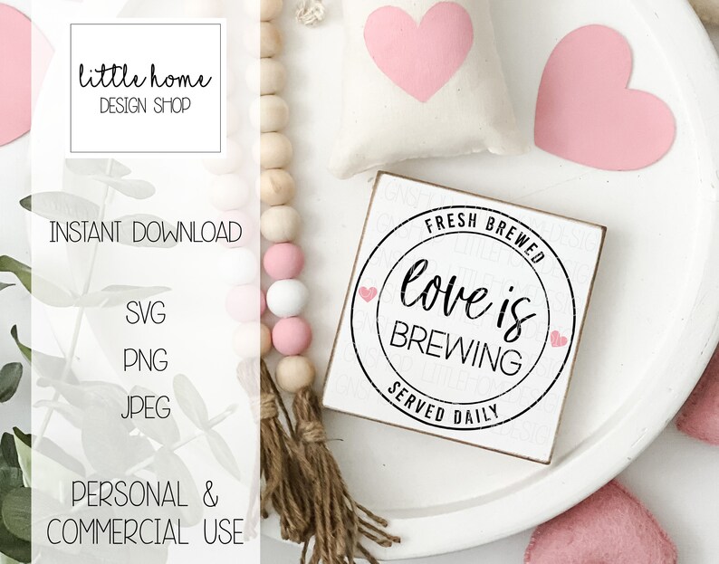 Op de afbeelding: Een wit houten bord met zwarte tekst die "Fresh Brewed Love is Brewing Served Daily" luidt. Het bord is omringd door roze harten en een houten kralensnoer.