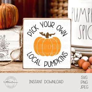 Puede incluir: Un letrero de madera blanco con el texto "Pick Your Own Local Pumpkins" en letras negras. Una calabaza naranja estilizada con un tallo marrón está en el centro del letrero.
