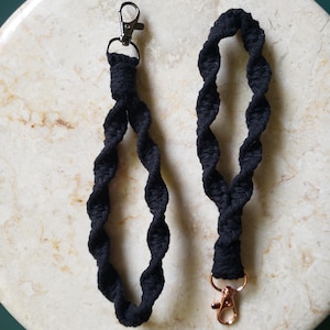 May include: Two black macrame wrist lanyards with metal swivel clasps. One has a silver clasp, and the other has a copper-colored clasp. The lanyards are made of twisted black cord and are displayed on a light-colored, speckled surface.
