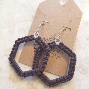 May include: A pair of dark brown macrame earrings with a geometric, square shape. The earrings hang from silver-colored hooks. They are displayed on a brown tag with a daisy design.