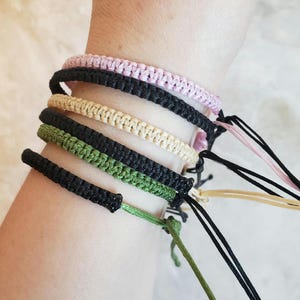 May include: A set of five friendship bracelets in black, green, beige, and pink. The bracelets are made of woven string and have adjustable closures.