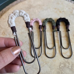 May include: Four large metal paper clips, each featuring a decorative top section wrapped in knotted cord. The colors of the cord include cream, light pink, olive green, and black, with dark brown thread accents securing the ends of the knots.