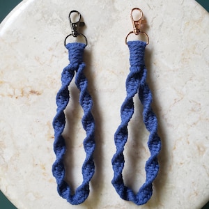 May include: Two blue macrame keychains with metal clasps. One keychain has a black clasp, and the other has a rose gold clasp. The keychains are made of twisted blue cord and are displayed on a light-colored, circular surface.