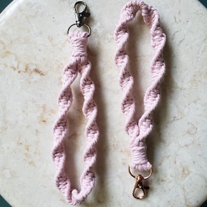 May include: Two pink macrame lanyards with metal clasps. One lanyard has a black clasp, and the other has a gold clasp. The lanyards are made of twisted, soft pink cord, and are displayed on a marble surface.