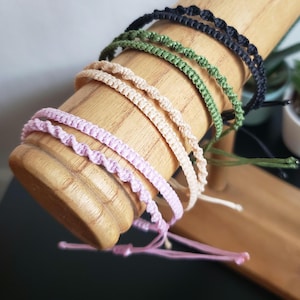 May include: A set of five colorful friendship bracelets in pink, beige, green, and black. The bracelets are made of thin, braided cord and are displayed on a wooden dowel.