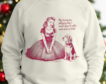 May include: A cream-coloured sweatshirt featuring a red graphic print of a person in a polka-dot dress kneeling beside a beagle. The text reads: My headache playing like I won't pop 10 pills and end us both. The design is centred on the chest.