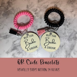 May include: Two pink and black silicone bracelets with a key ring attached to each. The key rings have round tags that say "Buy the Bride a Drink" in black script on a cream background.