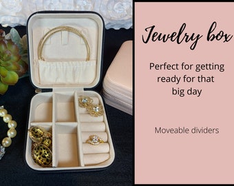 Pode incluir: Uma caixa de joias preta com interior bege e vários compartimentos. A caixa está aberta e contém um colar de ouro, brincos e anéis. O texto "Jewelry box" está escrito em itálico sobre um fundo rosa. O texto "Perfect for getting ready for that big day" está escrito abaixo do título. O texto "Moveable dividers" está escrito na parte inferior do fundo rosa.