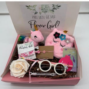 May include: A pink gift box with the text "Will You Be My Flower Girl?" contains a plush unicorn, sunglasses, a fluffy pink pouch, a notepad, a bracelet, hair ties, a rose, and a bubble wand. The box is filled with brown paper.