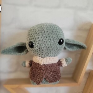 Peut inclure: Un jouet en peluche vert et marron crocheté ressemblant à un personnage de la franchise Star Wars. Le jouet a de grands yeux et une petite bouche.