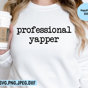 以下が含まれることがあります： 白いスウェットシャツに黒い文字で「professional yapper」と書いてあります。文字はタイプライターのフォントです。スウェットシャツは、コーヒーカップを持っている人が着ています。
