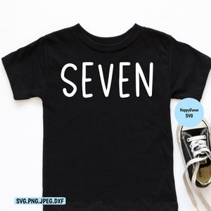 May include: Black t-shirt with the word "SEVEN" printed in white. The shirt is laying flat on a white surface. There are two black and white sneakers on the right side of the image.
