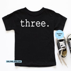 May include: Black t-shirt with the word "three" printed in white on the front. The shirt is laying flat on a white surface. There are two black and white sneakers on the right side of the image.