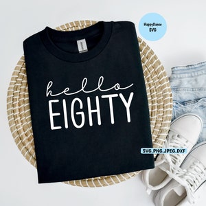 May include: Black t-shirt with white text that says "hello eighty" on a woven basket with a pair of white sneakers and blue denim shorts.