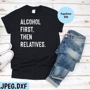 Puede incluir: Camiseta negra con texto blanco que dice "Alcohol First, Then Relatives." La camiseta está colocada sobre una superficie de madera blanca con un par de zapatillas blancas y unos vaqueros azules.