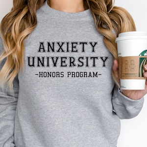 May include: A light gray sweatshirt with the text "ANXIETY UNIVERSITY - HONORS PROGRAM" in black block letters. The sweatshirt is long-sleeved and has a crew neck. A white coffee cup with a brown sleeve is held in the hand.