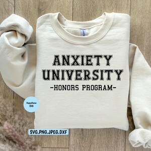 May include: A cream-colored sweatshirt with the text "ANXIETY UNIVERSITY - HONORS PROGRAM" in black block letters. The sweatshirt is laid flat on a light wood surface. A small blue circle with text is in the lower left corner.