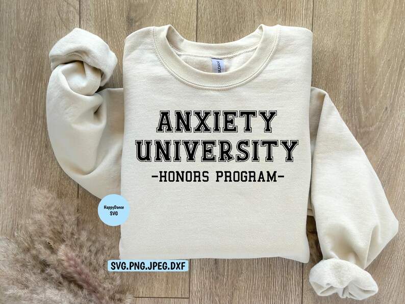 May include: A cream-colored sweatshirt with the text "ANXIETY UNIVERSITY - HONORS PROGRAM" in black block letters. The sweatshirt is laid flat on a light wood surface. A small blue circle with text is in the lower left corner.