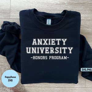 May include: A navy blue sweatshirt with the text "ANXIETY UNIVERSITY - HONORS PROGRAM" in white block letters. The sweatshirt is folded on a light wood surface. A small blue circle with the text "HappyDance SVG" is in the lower left corner.