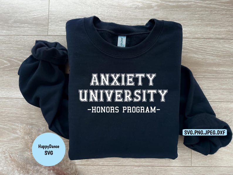 May include: A navy blue sweatshirt with the text "ANXIETY UNIVERSITY - HONORS PROGRAM" in white block letters. The sweatshirt is folded on a light wood surface. A small blue circle with the text "HappyDance SVG" is in the lower left corner.