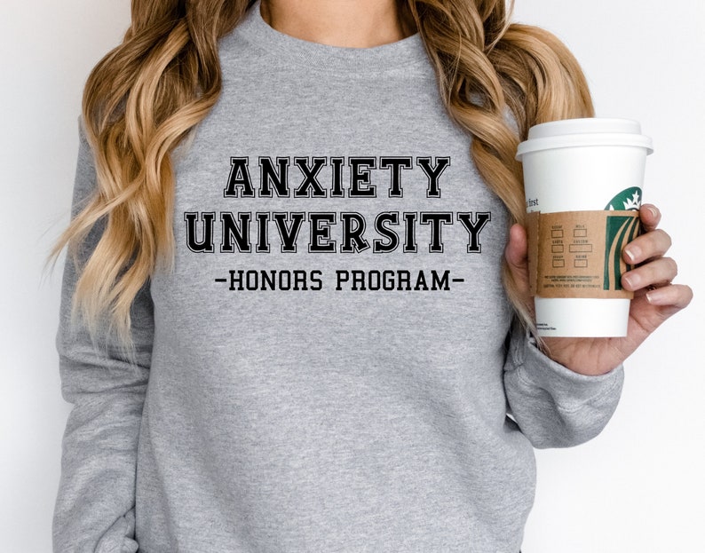 May include: A light gray sweatshirt with the text "ANXIETY UNIVERSITY - HONORS PROGRAM" in black block letters. The sweatshirt is long-sleeved and has a crew neck. A white coffee cup with a brown sleeve is held in the hand.