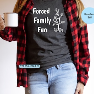 May include: Dark grey t-shirt with the text "Forced Family Fun" and a white graphic of a tree with baubles. The person is wearing a red and black checked shirt and blue jeans, holding a white mug.
