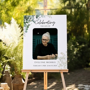 Pu&ograve; includere: Un cartello commemorativo bianco con una foto di una donna che indossa una camicia nera e occhiali. Il cartello &egrave; decorato con fogliame verde e il testo "Celebrating the life of Evelyne Morris February 1, 1968 - June 10, 2024".