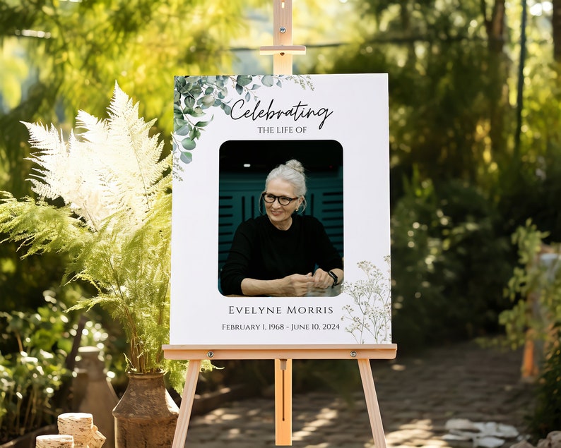 Pu&ograve; includere: Un cartello commemorativo bianco con una foto di una donna che indossa una camicia nera e occhiali. Il cartello &egrave; decorato con fogliame verde e il testo "Celebrating the life of Evelyne Morris February 1, 1968 - June 10, 2024".