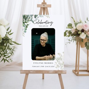 Pu&ograve; includere: Un cartello commemorativo bianco con un bordo floreale verde e una foto di una donna. Il cartello recita "Celebrating the Life of Evelyne Morris February 1, 1968 - June 10, 2024".