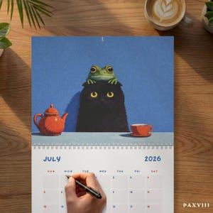 May include: A July 2026 wall calendar page featuring an illustration of a black cat with a green frog on its head, sitting at a table with an orange teapot and teacup against a blue background. The calendar grid shows the month and year.