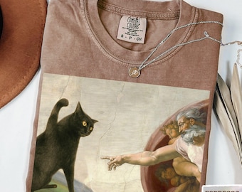 May include: A brown crew neck t-shirt featuring a graphic print of a black cat reaching toward a hand in a style reminiscent of The Creation of Adam. The shirt has a Comfort Colors label inside the collar and a silver-toned necklace rests on top.